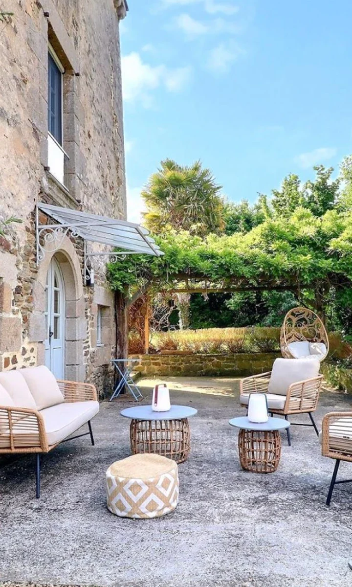 Photographie d’une terrasse de gîte en pierre, salon en rotin sous une pergola.
